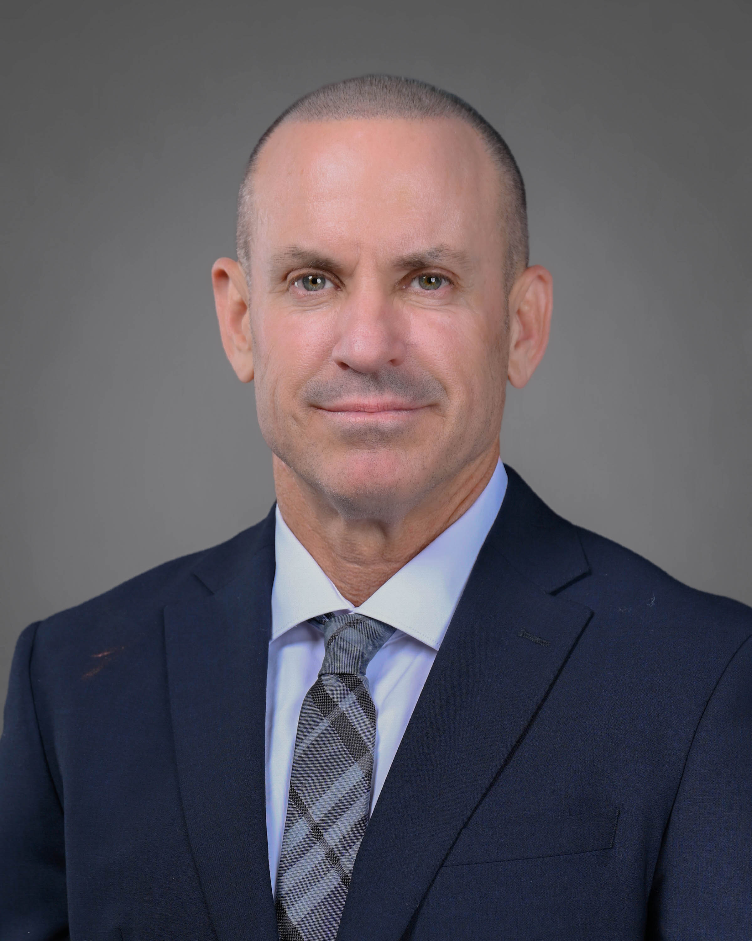 Clean cut and clean-shaven man in a navy blue suit with a lighter blue shirt and plaid tie.
